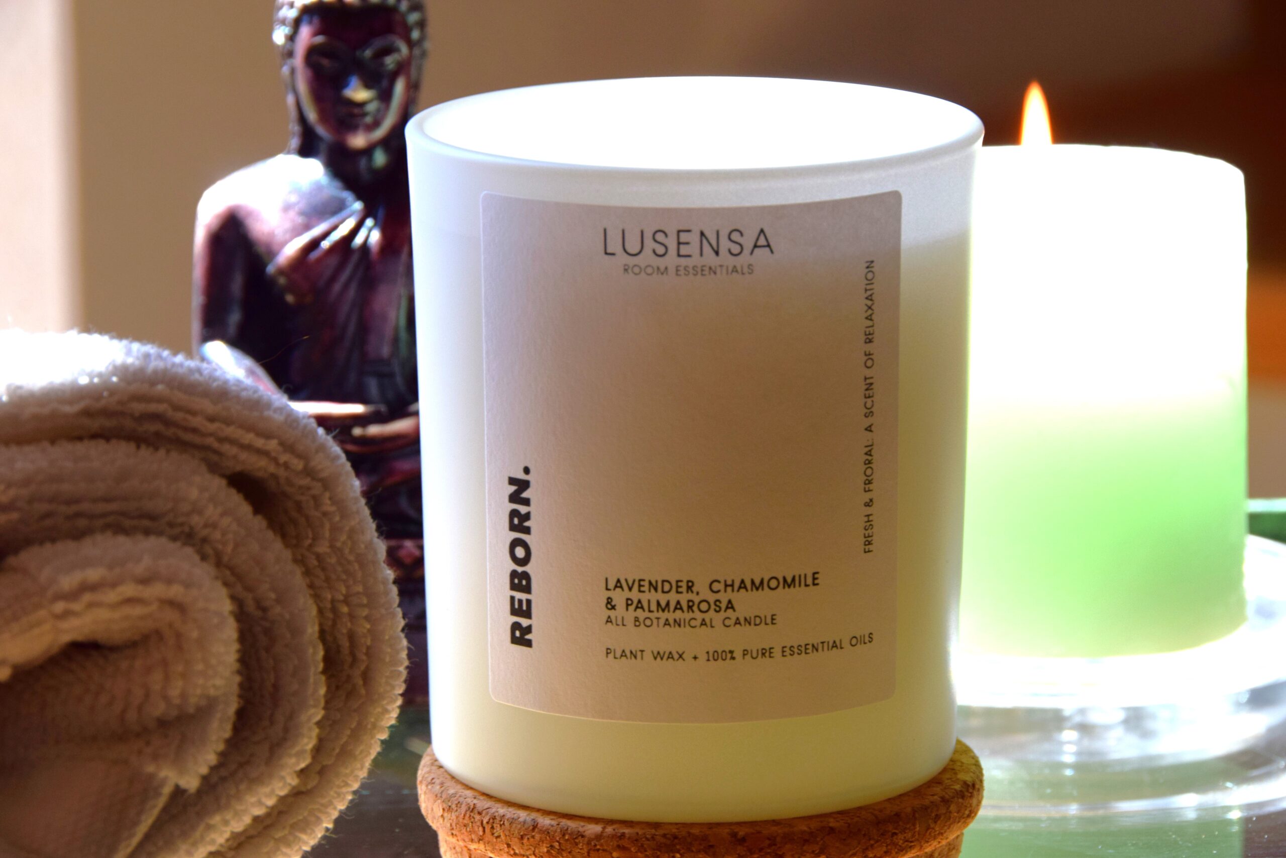 lusensa-duftkerze-reborn-mood Bild mit einer Duftkerze im Glas, eine brennende Kerze und einer Buddha-Figur im Hintergrund